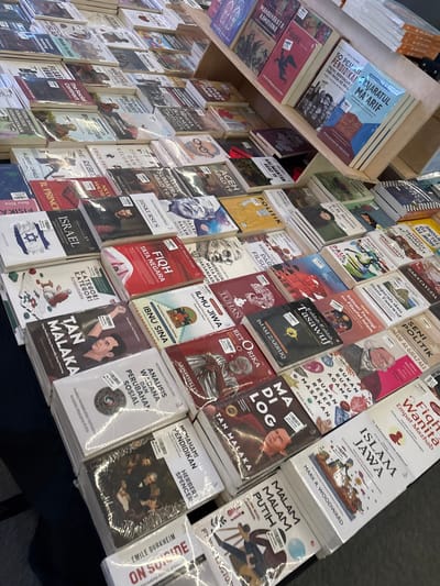 This media file features a close-up, slightly angled view of a bookshelf filled with books. The books are predominantly Indonesian titles, with some clearly visible in Bahasa Indonesia and others with Arabic script. The subjects of the books appear to be varied, including philosophy, religion, history, and politics, with titles like "Filsafat Timur," "Sejarah dan Ilmu Tasawuf," "Figh Wanita Empat Mazhab," "Islam Jawa," "Analisis Wacana dan Perubahan Sosial," and "Tan Malaka."

The books are neatly arranged on shelves, suggesting a bookstore or library setting. The lighting is bright, indicating an indoor environment, possibly during the daytime. The presence of plastic wrapping on some of the books suggests they are new or for sale. The overall impression is one of an organized collection of educational and cultural material. No specific people are clearly depicted, though some book covers feature illustrations or portraits of individuals. The setting is implied to be Wanasari, Indonesia, as per the provided location context.