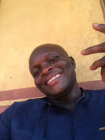 An adult dark-skinned male is captured in a close-up, slightly low-angle photograph, smiling broadly with white teeth exposed, and looking directly at the camera. He is bald or has extremely short dark hair and appears to be outdoors. He wears a dark blue long-sleeved garment, possibly a tunic, with a Mandarin collar fastened by snap buttons. A small, light-colored rectangular emblem, possibly reading "Fashion," is visible on the right chest area of his clothing. His left hand is raised to the upper right of the frame, with the index and middle fingers extended in a 'V' shape. The background consists of a textured, faded yellow or light orange wall with a horizontal dark red stripe running across its lower portion, behind the man's left shoulder and chest. The location is in the city of Ugwolawo, Nigeria.