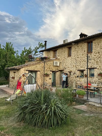 The image depicts a stone house with a tiled roof, likely in Cannara, Italy. A man in a light blue t-shirt and dark pants stands near an open doorway. A woman in a red dress is visible near a swing set with laundry hanging on it. In the background, another person in a red shirt is seated at a picnic table with other individuals. The environment includes a lawn, various plants, and a cloudy sky.