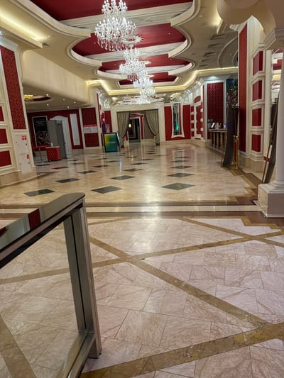 This image captures a grand, ornate interior hall, likely within a building in Shkotovo, Russia. The scene is devoid of people, focusing instead on the elaborate architecture and decor.
The dominant features are the richly decorated walls and ceilings, contrasting with a highly polished, reflective floor. The walls are predominantly a deep red with intricate patterned wallpaper or fabric, framed by elegant beige and white moldings, arches, and classical-style columns. Multiple large, sparkling crystal chandeliers hang from the arched ceilings, providing abundant artificial light that reflects off the glossy surfaces. Additional recessed lighting illuminates the arches and coves, enhancing the luxurious atmosphere.
The floor is a mosaic of marble or high-gloss tiles, featuring complex geometric patterns. It comprises various shades of beige, cream, and brown, with striking diagonal stripes and diamond shapes in darker hues. The reflective quality of the floor creates mirror-like reflections of the chandeliers and architectural elements above.
Visible objects include a modern digital display stand with the green and yellow "СБЕР" (SBER) logo, indicating the presence of a Sberbank service or advertisement. Further along the hall, a poster with "ШВЕДСКАЯ ЛИНИЯ" (Swedish Line) and "Baxi" is discernible. A fire extinguisher sign is also visible on a wall. In the immediate foreground, part of a glass barrier or partition is visible, framing the lower right corner of the shot.
The lighting suggests an indoor setting, likely in the evening or a space where natural light is not the primary illumination. The overall impression is one of opulence, formality, and grandeur, reminiscent of a luxury hotel, theater lobby, or upscale commercial complex.