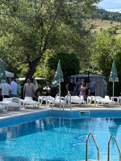 In Lozno, Bulgaria, this image captures an outdoor gathering around a swimming pool on a sunny day. A group of people, dressed in casual and semi-formal attire, are standing near the pool. To the left, two men and a woman in a green dress are conversing. A man in a light-colored shirt stands slightly apart, facing forward. In the background, several other individuals are gathered near a black metal fence, with some under green umbrellas that bear logos. White lounge chairs are arranged on the tiled deck surrounding the clear blue swimming pool, which has a mosaic pattern at the bottom. Lush green trees and foliage frame the scene, with a grassy hillside visible in the distance under a clear sky. A white vehicle is parked behind the group in the background.