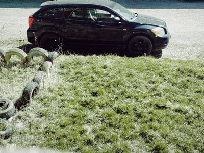 The scene in Klenovik, Bulgaria, depicts an outdoor environment under bright daylight conditions. A black five-door hatchback vehicle, consistent with a Dodge Caliber, is parked on a light-colored, possibly gravel or paved, surface in the upper-right quadrant of the frame. The vehicle features black wheels and a rear spoiler. A red and white striped object is partially visible inside the vehicle's rear driver-side window. In the foreground and extending along the left side, a patch of green and dry grass is present. This grassy area is bordered by a serpentine line of at least seven weathered, dark-colored vehicle tires, partially buried vertically in the ground. No individuals are present in the image.