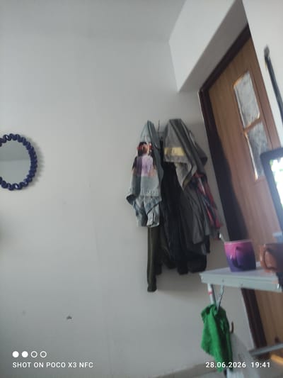 In a room with white walls, a blue, circular mirror with a decorative edge hangs on the left. To the right of the mirror, several pieces of clothing are hung on a rack, including a denim jacket with a colorful graphic on the back, a gray jacket, and other garments. A doorway with a wooden door and a glass pane is visible on the right side of the image. In the foreground, a white metal table is set up, holding a purple mug and a brown mug. A green cloth is hanging from the table. The timestamp "28.06.2026 19:41" is displayed in the bottom right corner, along with the text "SHOT ON POCO X3 NFC". The scene appears to be indoors, likely in a residential setting.