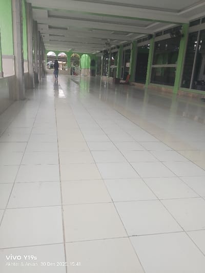 The image displays a long, well-maintained indoor corridor in Jagapura Lor, Indonesia, with a dominant light green and white color scheme. The floor consists of reflective white square tiles. A series of light green pillars with white bases and capitals line the left side of the corridor. The right side is characterized by large windows with green frames and a lower brown-colored section, allowing natural light into the space. The white ceiling features recessed lighting panels and two visible ceiling fans.
In the background, a single individual, dressed in dark clothing and a head covering, is observed walking away from the viewer. This person carries a light-colored bag or backpack, and their reflection is visible on the polished floor tiles. Along the right wall, in the middle distance, a reddish-brown object, consistent with a folded mat or small seat, rests against the wall. The far end of the corridor opens to an area with distinct arched doorways and visible outdoor greenery. The image includes a visible timestamp and device information at the bottom left: "vivo Y19s Akthar & Arvan 30 Des 2025, 15.41."