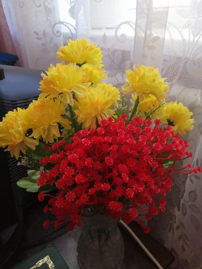 The image depicts a floral arrangement, likely artificial, positioned on a surface in front of a window with translucent curtains. The bouquet is composed of bright yellow, chrysanthemum-like flowers and a mass of small, vibrant red flowers, possibly baby's breath or similar filler. The yellow flowers are arranged in clusters, with some showing hints of orange at their petal tips. The red flowers form a dense, cloud-like mass at the base of the bouquet. The vase holding the flowers appears to be a textured, greyish-brown material. In the background, the curtains have a floral pattern that is partially obscured by the brightness of the light filtering through. To the left, a dark, possibly upholstered object is partially visible. In the lower left corner, a corner of a dark green book with ornate gold detailing is visible. The overall setting suggests an indoor environment, possibly a home or office, with a focus on decorative elements. The location of Lanivtsi, Ukraine, is not directly discernible from the visual elements in the image.