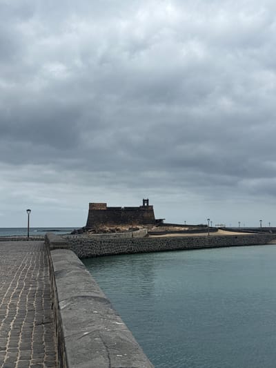 The image depicts a seawall in Arrecife, Spain, under a cloudy sky. The Castillo de San Gabriel, a stone fortress, stands next to the water. The water itself is calm with faint ripples. A paved walkway extends from the bottom right corner towards the horizon, lined with lamp posts. Another seawall structure is visible in the distance. The scene appears to be deserted, without any visible human activity.