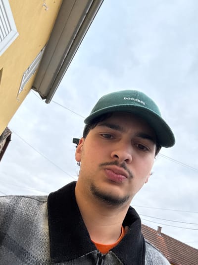 A young adult male is depicted in a close-up, upward-angled outdoor photograph, taken in Holtzheim, France. He faces the camera directly with his lips slightly pursed. The subject has dark hair, a light mustache, and stubble. He wears a dark green corduroy baseball cap with "DOCKERS" embroidered in white, an orange t-shirt, and a grey and black patterned jacket featuring a black collar and visible zipper. A small earring is present in his left ear. The background consists of an overcast sky. To the upper left, a bright yellow building with white window frames and a grey rain gutter is partially visible. Thin utility wires traverse the sky, and a reddish-brown tiled roof is partially visible in the lower right.