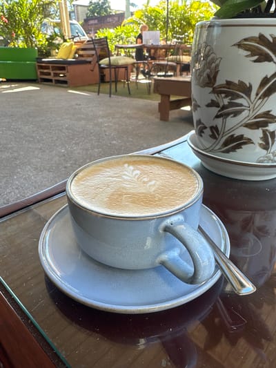 A close-up shot captures a light blue ceramic mug of latte art resting on a matching saucer. A silver spoon lies across the saucer. The mug and saucer are positioned on a dark, reflective glass-topped table. To the right, a portion of a ceramic pot decorated with brown floral patterns is visible. In the background, the scene opens to an outdoor seating area of a cafe, with various tables and chairs. Sunlight filters through lush green foliage, suggesting a bright day. A glimpse of a person can be seen in the distance, seated at a table. The environment suggests a relaxed cafe setting, likely in Bellingen, Australia, given the implied presence of outdoor seating and lush greenery characteristic of the region.