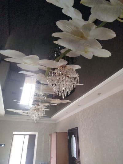 A dimly lit room features a dark, glossy ceiling adorned with a printed image of white orchids and several crystal chandeliers. The orchids appear to be large and stylized, with visible petals and stems. The chandeliers are ornate, with numerous dangling crystals that reflect light. One larger chandelier is prominently displayed in the center of the image, while two smaller ones are visible further down and to the left.
The walls of the room are covered in a textured, light-colored wallpaper. The ceiling transitions to a white crown molding, which is illuminated by recessed lighting fixtures. In the lower portion of the image, a doorway with a dark wooden frame and a glass inset is visible on the right. To the left of the doorway, a portion of a light-colored wardrobe or cabinet is visible. A large window, letting in bright daylight, is seen on the left side of the room. The overall impression is one of a modern, somewhat opulent interior. The location is identified as Жаңы-Айыл, Kyrgyzstan.