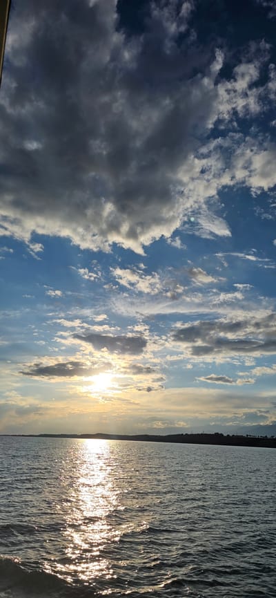 A wide shot shows a body of water reflecting the sun and sky at dusk. The sky is filled with dramatic, dark clouds interspersed with patches of bright blue and scattered white clouds. The sun is partially obscured by clouds, casting a golden light across the water, creating a shimmering reflection. In the distance, a dark silhouette of a treeline and mountains marks the horizon. The water is calm with a slight ripple. The scene appears to be a natural landscape, possibly a lake or a wide river, under a partly cloudy sky. There are no people or man-made structures visible in the foreground or midground. The lighting suggests the time of day is either sunrise or sunset.