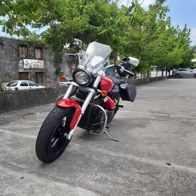 A red and black cruiser motorcycle is parked on a concrete surface, possibly a parking lot. The motorcycle is positioned at a slight angle, facing towards the right of the frame. It features a large, clear windshield, chrome accents on the engine and handlebars, and a black saddlebag attached to the rear. In the background, a stone wall with wooden window frames and a faded poster is visible. Trees with lush green foliage are overhead and to the right, with a blue sky peeking through. Further in the background, a white car is parked near the wall, and a modern gray car is seen in a parking area. The overall environment appears to be an urban setting in Vila Verde, Portugal. The scene is static and there are no people present.