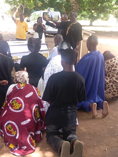 The image shows an outdoor gathering in what appears to be Ninzo South, Nigeria. A group of people is gathered around a white coffin. Several individuals are kneeling on the ground. One man dressed in a black robe is gesturing with his hand. Another man in a yellow shirt is standing behind the coffin with his arm raised. A woman is holding up a phone, seemingly taking a photo. An ambulance is parked in the background. The gathering takes place on bare ground with trees in the background. Some attendees are wearing clothes that have the words "CHRISTIAN WOMEN FELLOWSHIP" and "GARRISON" printed on the clothes.