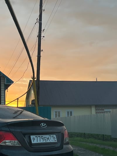 The image captures a twilight scene in the city of Bol'shoy Kukmor, Russia. In the foreground, the rear of a dark-colored car is visible, displaying a license plate that reads "P779EM 716 RUS". Behind the car, a grey corrugated metal fence stretches across the frame, with greenery and a dirt path running alongside it. Beyond the fence, a pale yellow house with white window frames is partially visible. A dark grey roof of another structure is prominent in the mid-ground. Overhead, a wooden utility pole with electrical wires cuts across the sky, which is filled with an orange and grey gradient of clouds, suggesting either sunrise or sunset.