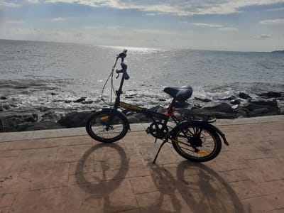 A black folding bicycle with a red lock is parked on a tiled walkway next to a rocky shoreline. The sea stretches to the horizon under a cloudy sky. The sun reflects off the water, creating a bright glare. The walkway is made of large, reddish-brown tiles arranged in a grid pattern. The bicycle casts a long shadow on the walkway. In the distance, faint shapes of ships can be seen on the water. The scene appears to be a promenade or coastal path, likely in Atakum, Turkey, given the context.