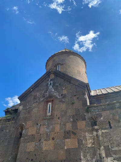 This media file is a low-angle, tilted shot of a section of an ancient stone building under a bright blue sky with scattered white clouds. The building appears to be constructed from large, irregular blocks of dark stone, interspersed with some lighter, more square-cut stones. Architectural details visible include a rounded tower with a narrow, arched window and a steeply sloped section of the main building with another long, narrow window framed by decorative stonework. Small green plants are growing in crevices in the stone walls, suggesting the building is old and exposed to the elements.
The lighting suggests it is daytime, likely mid-morning or early afternoon, given the brightness of the sky and the absence of strong shadows. The weather appears clear and fair. The perspective emphasizes the scale and texture of the stone structure against the vastness of the sky. The tilted angle gives a dynamic and dramatic feel to the scene.
Based on the provided location context, this is likely a view of a church or monastery in Saghmosavan, Armenia, known for its ancient stone architecture. No people are visible in the frame, and there is no discernible text. The scene evokes a sense of history, durability, and architectural grandeur.