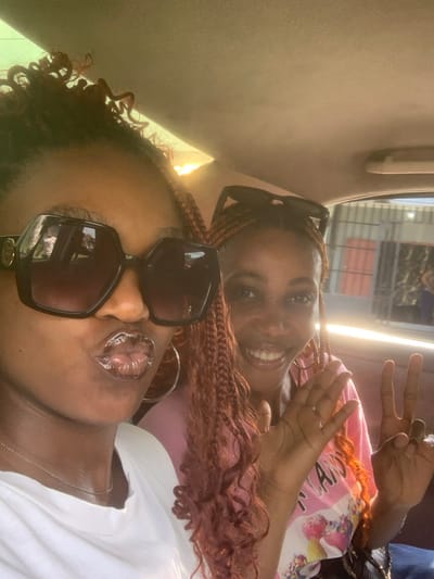 The image displays two adult females inside a vehicle located in Anantigha, Nigeria. The woman on the left, in the foreground, has dark skin, dark curly hair, and wears large, octagonal dark sunglasses with a gold accent. She is wearing a white t-shirt and a thin silver-colored chain, with her lips pursed in a glossy "kissing" pose. A large silver-colored hoop earring is visible on her left ear. The woman on the right, positioned slightly behind and to the right, has a lighter complexion, reddish-brown braided hair with dark sunglasses resting on her head. She is smiling broadly and making a "peace" sign with her left hand, while her right hand is slightly raised. She wears a light pink t-shirt with a partial graphic that includes the word "CANDY". The vehicle's interior features a light-colored, possibly beige, headliner. Sunlight is visible entering from the upper left. Through the vehicle's windows, blurred urban elements are visible, including what appears to be a corrugated metal barrier and part of a building with a red-colored section.