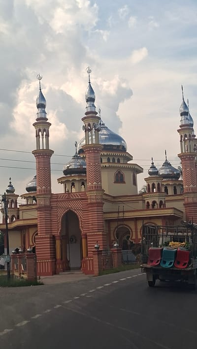 A mosque with a distinctive red and yellow facade and silver domes is visible on a cloudy day. The mosque has tall minarets topped with crescents. In the foreground, a truck with three colorful plastic chairs is parked on the side of a road. The road has visible white lane markings. Some greenery is present to the left of the mosque. The image appears to be taken in Cempa, Indonesia, based on the presence of the mosque which is a notable landmark in that area.