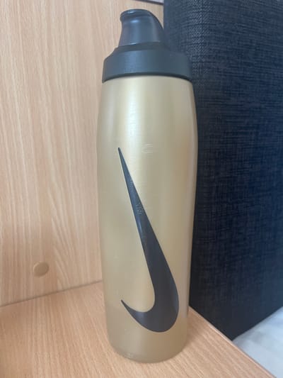 A gold-colored Nike water bottle with a black Nike swoosh logo is standing upright on a light-colored wooden surface. The bottle has a black cap and appears to be made of plastic. Behind the bottle, a dark textured fabric is visible, which could be part of a sofa or chair. The background on the left is a light-colored wooden wall with visible grain. There is a small, round, light-colored object on the wooden wall to the left of the bottle, possibly a knot in the wood or a small decorative element. The lighting suggests an indoor environment. No people are present. The scene is indoors and does not provide specific details about the city of Toila, Estonia.