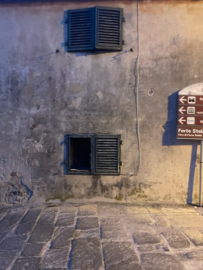 The image shows a section of an exterior wall in Portoferraio, Italy. The wall is made of aged, light grey plaster and has two windows with dark green wooden shutters. The upper window is closed, while the lower one is partially open. The wall has a visible crack running down the center. A sign is attached to the wall, with three arrows pointing left and listing directions in Italian. There is a paved, cobblestone walkway in front of the wall.  The image is shot at a low angle, with the wall taking up most of the space.