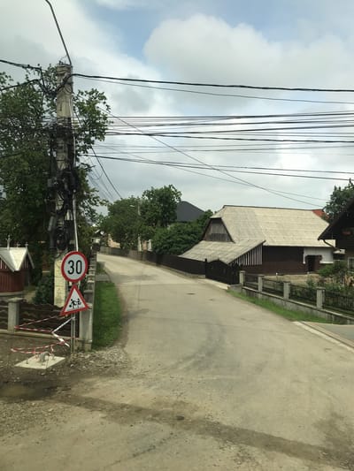 This image depicts a residential street scene in Vicovu de Sus, Romania, under a partly cloudy sky. The foreground features a dirt and gravel shoulder transitioning to a paved asphalt road that ascends gently and curves to the right, disappearing into the distance. On the left side, a large concrete utility pole is heavily burdened with numerous black electrical and communication cables, some coiled and bundled. Attached to a smaller post beside the utility pole are two road signs: a circular speed limit sign indicating "30" and a triangular warning sign depicting two children, suggesting a children crossing zone. At the base of these signs, a concrete slab is surrounded by red and white striped warning tape on the rough ground, which includes a small puddle. Further left, a small structure with a white roof and red base is partially visible. The road's right edge is bordered by a concrete curb, a narrow strip of grass, and a continuous fence constructed from concrete posts and black metal panels. Beyond this fence, a house with a large, light grey, corrugated roof and dark brown walls is prominent, featuring a distinct architectural style. Another building with a dark tiled roof is visible further back. Green trees and dense foliage are present on both sides of the road and in the background. No people or vehicles are visible.