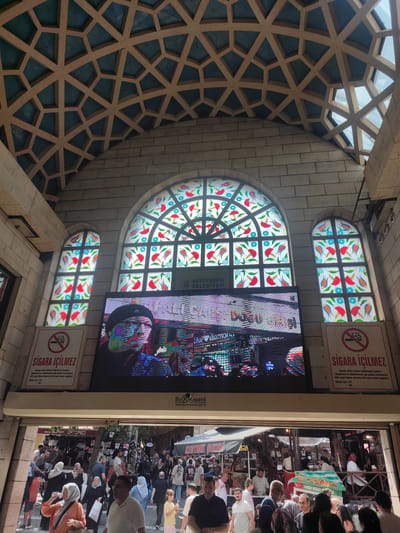 The image captures a view looking upwards and into a marketplace in Osmangazi, Turkey. The architecture above features a distinctive geometric ceiling design composed of light-colored beams forming hexagonal and triangular shapes, with a dark blue material filling the spaces. Below this ceiling, a brick wall is visible, supporting two large stained-glass windows. The windows are arched at the top and feature intricate patterns of red, blue, and orange floral designs. Between the windows, a large digital screen displays a vibrant, pixelated image of a person wearing a black hat, along with text and graphics that are largely illegible due to the image's resolution and the screen's display. Flanking the digital screen are two white signs with red lettering and a red circle with a diagonal line through a cigarette symbol, indicating a smoking prohibition. The signs are in Turkish, with the prominent text "SIGARA İÇİLMEZ" (No Smoking).

Below the screen and signs, an open entrance leads into the marketplace. A crowd of people are visible within the marketplace, some browsing stalls that appear to sell footwear and other goods. The lighting suggests it is daytime, with sunlight filtering through the ceiling and windows, creating some glare. The overall impression is of a busy, covered public space with decorative architectural elements.