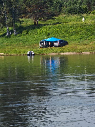 In Podporozh'e, Russia, on a sunny day, a group of people are gathered on the grassy bank of a body of water, possibly a river. A blue tarp is stretched over a picnic area, sheltering two vehicles, one a dark SUV and the other a white van. A person in blue shorts stands near the van. A small inflatable boat with an outboard motor is partially submerged in the water, close to the bank. The environment is characterized by lush green grass, trees, and the rippling surface of the water, which reflects the blue tarp and the surrounding landscape. The scene suggests a recreational activity, such as camping or fishing, taking place in a natural setting.