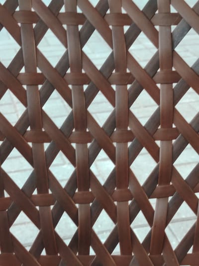 This is a close-up, high-angle shot of a woven brown wicker material, possibly a chair or fence, set against a blurred background of what appears to be a light-colored tiled surface. The wicker is composed of interlocking strips forming a diamond pattern with diamond-shaped openings. The lighting is even, and the focus is sharp on the texture and weave of the material, with the background appearing out of focus. There are no people or animals visible in the image, and no discernible geographical markers to suggest a specific location in Toranghylyq, Kazakhstan. The image appears to be a detail shot focusing solely on the material's pattern.