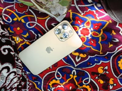 A light gold iPhone 13 Pro is positioned diagonally on top of a vibrantly colored, patterned fabric. The fabric features a complex design with floral motifs in shades of red, blue, yellow, and deep purple, outlined in white. The iPhone's rear camera array, with its three lenses, is visible on the upper right side of the device. The Apple logo is subtly embossed on the back of the phone, towards the center-left. Part of a dark gray object, possibly clothing or a bag, is visible in the upper right corner of the image, casting a shadow on the fabric. In the upper portion of the image, a translucent white material with delicate white floral patterns overlays the colorful fabric. The scene is illuminated, with reflections visible in the iPhone's lenses. The location is identified as Osh, Kyrgyzstan, suggesting this image was taken in a setting that incorporates traditional Kyrgyz textiles or decor.