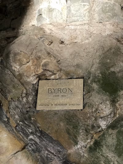 The image captures a close-up view of a stone plaque mounted on a rough, uneven rock wall. The plaque commemorates Lord Byron, inscribed with "BYRON JUIN 1816" and "1788 t 1824 IN MEMORIAM 27 MAI 1924." The stone has a light gray color with darkened edges and some signs of weathering. The surrounding rock wall exhibits a mix of light and dark gray hues, with visible cracks, crevices, and irregular textures. There are possible signs of moisture or moss in some areas of the rock wall. This location is likely a memorial at the Chateau de Chillon, in Veytaux, Switzerland.