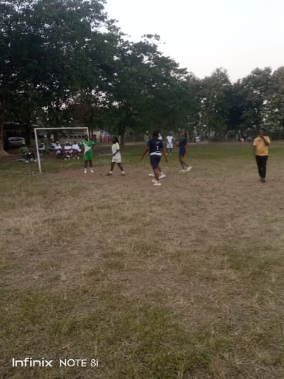 A group of people are playing a game on a grassy field. Some are actively participating while others are sitting on the sidelines near a goalpost. There is a man in a yellow shirt walking on the right side of the image. Trees are present in the background and the sky is overcast. The image has a smartphone watermark at the bottom.