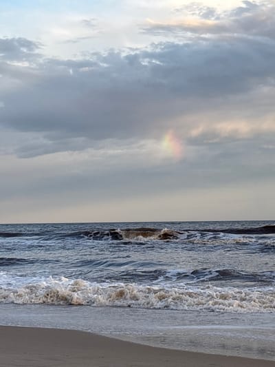 The image shows a beach scene divided roughly in half. The left side displays a cloudy sky with a faint rainbow-like band of color. The right side shows the ocean with waves crashing onto the sand. The water is a muted blue-gray, and the sand is a light beige. The picture is oriented vertically.