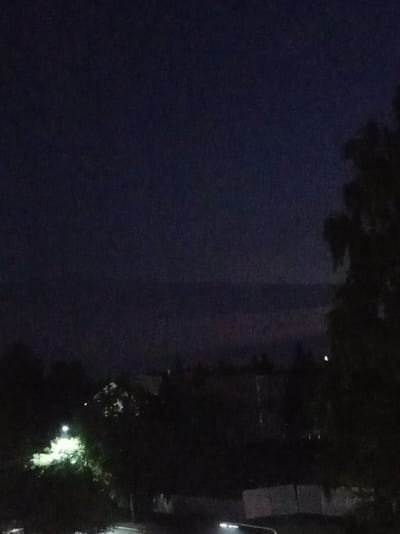 The image is a nighttime landscape shot, possibly taken from an elevated position. The sky is dark blue with a hazy band across the horizon, suggesting either clouds or distant lights. Silhouetted trees and buildings are visible in the foreground and midground. A bright street light illuminates some foliage to the left of the frame. The overall impression is of a suburban or semi-rural area at night. Without additional information or visual cues, it is impossible to determine the specific location within Alatyr, Russia.