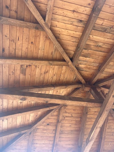 The image displays the underside of a wooden roof structure, situated in Gigintsi, Bulgaria. The ceiling surface consists of numerous light-to-medium brown wooden planks, exhibiting natural grain patterns and knots. A structural framework of darker brown, rectangular wooden beams provides support, with multiple diagonal beams converging towards a central, multi-faceted wooden block located in the lower-middle area of the frame. Additional beams run horizontally and vertically relative to the frame edges. The wood shows some minor, localized discoloration and staining. The scene is illuminated by natural light, originating from below and the lower-left, casting subtle shadows. No individuals, objects beyond the architectural elements, or actions are present.