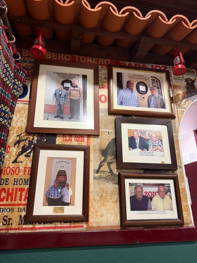 The image displays an interior wall of a traditional establishment, likely a tavern or restaurant, located in Santarém, Portugal. The upper portion of the scene reveals an exposed ceiling featuring rows of reddish-brown terracotta roof tiles and dark wooden beams, from which two red, vintage-style oil lanterns are suspended.
The central focus is a collection of five framed photographs, each encased in a dark brown wooden frame with light matting, mounted on a textured wall largely covered by overlapping, aged bullfighting posters. Visible poster fragments include "BOBERO TOREO", "15,30 horas", "JOAO MOURA", and "PLAZA DE TOROS DE". A silhouette of a charging bull is discernible between some frames.
The photographs depict groups of older men, indicative of camaraderie or commemoration, often associated with "Taberna do Quinzena" as suggested by text on or near the photos:
1. **Top Left (Vertical)**: Two standing men, one in a light suit holding a cane, the other in a beige sweater. Inscription mentions "António Manhosa e Mário Pereira" and "15. Novembro. 2008".
2. **Top Right (Horizontal)**: Two men posing indoors, one in a striped shirt, the other in a light jacket. A circular "TABERNA DO QUINZENA" sign is visible behind them. Inscription mentions "Rui Ferrer e João Mário Bernardez" and "13. Out. 2007".
3. **Middle Right (Horizontal)**: Two men seated at a table, one in a dark suit, the other in a plaid shirt and glasses. Inscription mentions "Abel Reis e João da Paz" and "14.10.2006".
4. **Bottom Left (Vertical)**: Two standing men, one in a plaid shirt, the other in a white polo shirt. A drawing of a wooden barrel with "TABERNA DO" is above them. Inscription mentions "JOSÉ DO SEIXO - LUÍS CLEMENTE" and "16.10.2009".
5. **Bottom Right (Horizontal)**: Two smiling men, one in a purple sweater, the other in a yellow polo shirt, both wearing glasses. Inscription mentions "TOKALON e TANU" and "30. Out. 2010".
To the far left, a vertically hanging, multicolored, patterned fabric, possibly a textile or part of a decorative curtain, is partially visible. The lower section of the wall below the posters is painted a solid, dark green.