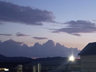 This image captures a twilight sky over the Баянауыл ауылдық округі in Kazakhstan. The sky transitions from a pale blue at the top to a softer gradient of pink and lavender near the horizon, where dramatic, bulbous clouds are silhouetted against the fading light. A small, bright green light is visible in the upper left portion of the sky, possibly a distant aircraft or star. Below the clouds, a dark, hilly landscape forms the horizon. In the foreground, the roofs of several buildings are visible, with one building on the right featuring a dark, peaked roof. A bright light source, likely a streetlamp or strong light fixture, emanates a sunburst effect from behind some structures in the mid-ground, casting light onto the surrounding buildings and landscape. The overall impression is one of a rural or semi-rural setting at dusk.