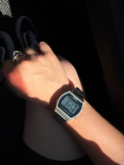 The image shows a close-up of a person's left arm wearing a silver digital Casio watch with a black metal band. The watch face displays the time as 9:47 and the date as 6-20. The person is also wearing a silver ring with an intricate design on their index finger. The arm is illuminated by a strong orange light, casting a shadow across it. In the background, partially visible, are the tops of black Vans sneakers. The scene appears to be indoors, possibly in a vehicle, given the dark surroundings and the texture of what might be upholstery. The location of Tsamakaberd, Armenia is not discernible from the visual information present in the image.