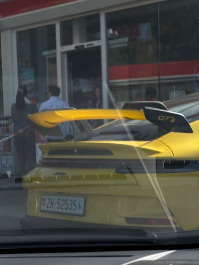 The image displays the rear of a bright yellow Porsche 911 GT3 sports car in Meuspath, Germany. The vehicle is equipped with a prominent black-accented swan-neck rear wing, displaying "GT3" on its right endplate. A Swiss license plate, "ZH-52535", is affixed to the rear bumper. The scene is observed through a reflective surface, indicated by diagonal light streaks across the frame.
In the background, a commercial building with large glass windows and a red overhead fascia is visible. An automatic sliding glass door serves as the primary entrance. The interior of the building reveals shelving with various items. Three individuals are identifiable near the entrance: a female in a striped top and dark pants, viewed from the back, positioned to the left of the doorway; a male in a light blue collared shirt and dark trousers, also viewed from the back, standing centrally near the entrance; and a third female figure partially visible through the glass inside the building to the right. A red and white chain barrier is present in the lower left foreground, adjacent to the building.