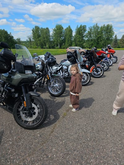 A line of motorcycles is parked on an asphalt surface next to a grassy area with trees. A young child wearing a brown sweatsuit stands in front of the motorcycles. A woman's legs are visible at the top of the image. The location is Borodinskogo Muzeya, Russia.