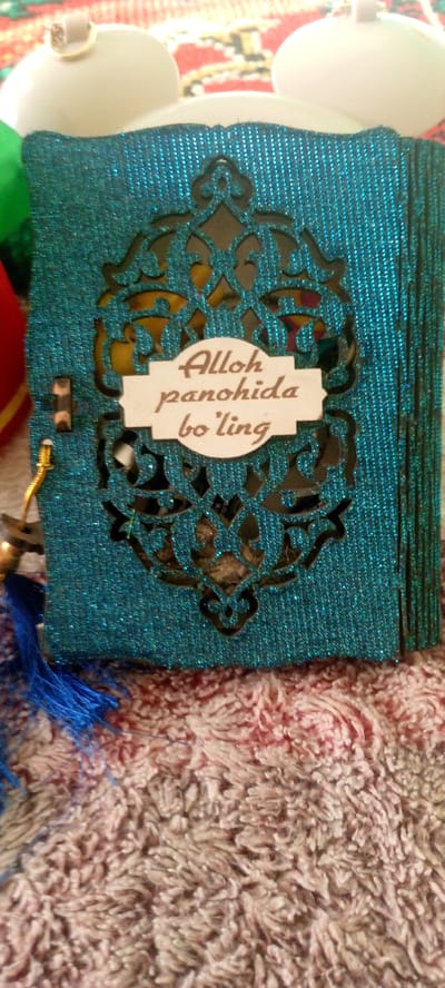 The image shows a close-up view of a turquoise, glitter-covered, decorative box with a cutout ornamental design on the front. Centered on the ornamental design is a small, light-colored label with dark text that reads "Alloh panohida bo'ling". A blue tassel and a golden string are attached to the left side of the box, near the bottom. The box rests on a textured, fuzzy surface, possibly a rug or blanket, with predominantly pink and white fibers. In the upper background, out of focus, are two white, round objects, possibly decorative ornaments or parts of a larger item, positioned behind the box. The overall presentation suggests the item is a gift or a decorative piece. There are no people or animals in the image. The location in Marhamat, Uzbekistan is not discernible from the visual content of the image.