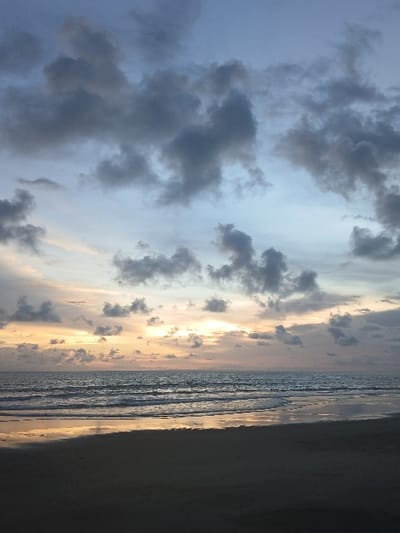The image depicts a coastal scene in Tibubiyu, Indonesia, under twilight conditions, likely sunrise or sunset. The foreground consists of dark, wet sand reflecting the sky's light. The midground features the ocean with multiple parallel waves breaking towards the shore. The water's surface reflects hues from the sky. The background is dominated by a cloudy sky, transitioning from dark grey and blue tones in the upper portion to lighter yellow, orange, and pink hues near the horizon, where the sun's light is visible through or behind scattered clouds. No individuals or man-made objects are visible within the frame.