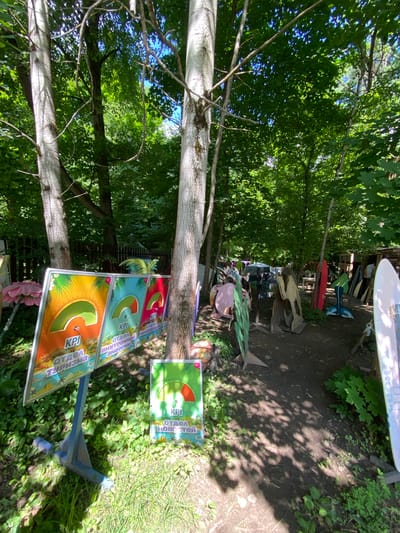 This media file captures an outdoor scene in Obninsk, Russia, likely during the daytime on a sunny day, indicated by the dappled sunlight and shadows on the ground. The setting appears to be a wooded area or yard, with mature trees and lush green foliage dominating the frame. Scattered among the greenery are various objects, suggesting a display or perhaps a workshop area.
Prominently featured are several colorful signs or posters, each featuring a gauge-like graphic with the letters "KPI" and some text in Russian. One sign reads "ОТДЕЛ НОВОСТЕЙ" (News Department), another "ОТДЕЛ ВНУТРЕННЕЙ ЭНЕРГИИ" (Internal Energy Department), and a third "ОТДЕЛ ТУРИЗМА" (Tourism Department). These signs are mounted on a blue metal stand.
In the background, other objects are visible, including what appears to be a surfboard or paddleboard, several pieces of what might be metal art or sculptures shaped like animals (such as a fish, a bird, and a whale tail), and some wooden planks. The overall impression is a somewhat eclectic and artistic arrangement in a natural setting. No people are clearly visible, though there might be a blurred figure in the distance behind some of the objects. The scene evokes a sense of a creative or recreational space, possibly an outdoor art installation or a place where outdoor activities are organized.