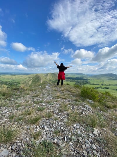 A person is standing on a rocky, grassy hilltop in Andreevka, Russia, with their arms outstretched and looking out at a vast green valley below. The sky is a bright blue with scattered white clouds. The person is wearing a black shirt and a red skirt. The terrain is hilly and covered in sparse vegetation and rocks. The scene appears to be a natural landscape with rolling hills and distant trees.