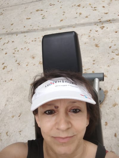 The image shows a woman wearing a white visor that reads "LUXEMBOURG LET'S MAKE IT HAPPEN". She is lying on what appears to be a park bench or similar outdoor seating, with her head tilted back. The background is a textured surface that looks like concrete with scattered debris, possibly leaves or dried grass. The lighting suggests it's daytime, and the overall mood is casual and relaxed. The woman has dark hair and is looking towards the camera with a slight smile. The setting appears to be outdoors, possibly in a park or recreational area within Vila Nova de Famalicão, Portugal.
