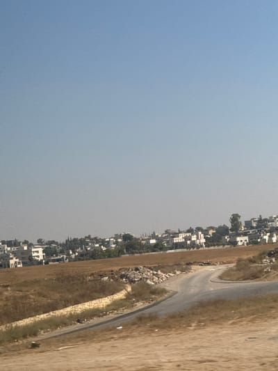 The image captures a landscape scene, seemingly a rural or semi-urban area, with a clear sky. On the left side of the frame, a vast expanse of pale blue sky dominates, transitioning to a lighter shade towards the horizon. To the right of the sky, a densely populated area with buildings is visible, stretching along a hillside. The buildings are primarily white and appear to be houses or apartments, with some greenery interspersed among them.

A winding road is visible in the middle ground, cutting through a dry, brownish landscape. The road is paved and appears to be in good condition, with a slight curve. The terrain surrounding the road is covered in dry grass and some scattered rocks.

Further to the right, the landscape continues with more dry, brownish terrain. There is a noticeable stone wall or embankment along the side of the road, constructed from roughly cut stones. There are a few small white specks on the road, which might be small stones or debris. A dark shape, possibly a person or a vehicle, is visible on the road, suggesting some form of activity or movement.

The overall impression is of a sunny day, likely in a warm climate, given the dry vegetation. The time of day appears to be daytime, with the sky being light and clear. The location is suggested to be the Bnei Shimon Regional Council in Israel. There are no visible signs or text in the image. The scene conveys a sense of everyday life in a developing or established community, with infrastructure like roads connecting it to the surrounding environment.