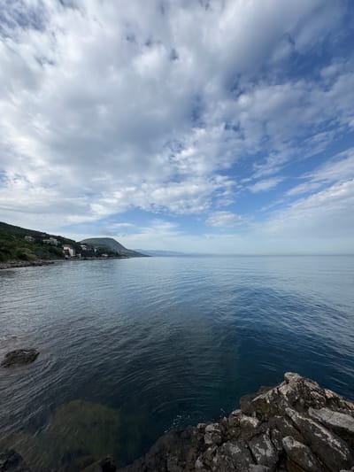 The image presents a view from what appears to be a coastal area, possibly in Bondarenkovo, Ukraine. The sky is predominantly light blue, interspersed with numerous white clouds. Below the sky, the sea stretches out, showing a slightly darker blue hue with subtle wave patterns. A landmass is visible in the midground with what appear to be buildings scattered along the coastal area. In the immediate foreground, there are rocks, suggesting a clifftop or rocky shoreline location. The overall composition indicates a scenic view, potentially capturing a moment in time from a high vantage point overlooking the sea and coastline.