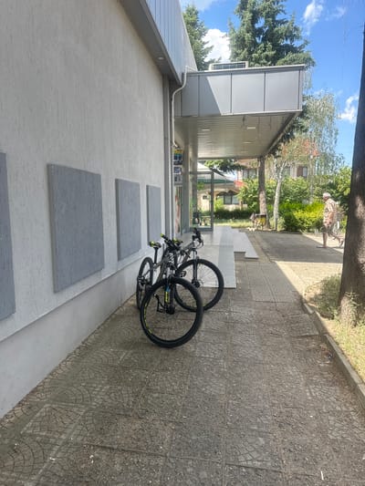 The image depicts an outdoor scene with two bicycles parked on a tiled pathway. The bicycles are dark-colored with yellow accents on the handlebars and rims. They are leaning against each other, positioned near the entrance of a building.
The building appears to be a commercial establishment with large glass windows and a light-colored facade. Several rectangular grey panels are mounted on the exterior wall of the building. Above the entrance, a white awning extends over the pathway.
In the background, there is lush greenery, including trees and shrubs, and residential buildings can be seen. A person, whose face is not clearly visible, is walking away from the camera on the pathway in the distance. The sky is blue with scattered white clouds, suggesting it is daytime. The sun casts shadows on the ground, indicating a bright, sunny day. The overall atmosphere seems relaxed and casual. The location is Ovoshtnik, Bulgaria. There is visible text on signage through the windows of the building, but it is not legible in this image.