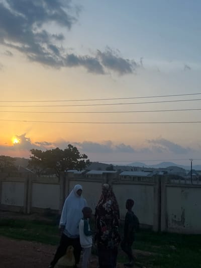 An outdoor scene in Rinjim Gani, Nigeria, is depicted at either sunrise or sunset, characterized by a bright, low-lying sun on the far left horizon. The sky exhibits a gradient from intense orange and yellow near the sun to a pale blue and light grey at the top right, with scattered dark and light clouds. Multiple parallel power lines traverse the upper-mid portion of the frame.
Four individuals, appearing as two adults and two children, are gathered on unpaved ground in the foreground. On the left, an adult wears a white head covering and light-colored garment over dark trousers, standing near a partially visible light-colored animal, possibly a chicken, on the ground. A young child stands in front of this adult, wearing a white and dark two-toned shirt and light trousers. To their right, another adult is dressed in a patterned, multi-colored garment that covers their head and body. To the right of this adult stands a second child, wearing dark clothing. All four individuals appear to be facing inwards towards the center of their group, engaged in an interaction.
In the mid-ground, a light-colored concrete perimeter wall runs horizontally behind the individuals. Behind this wall, various single-story buildings with light walls and dark roofs are visible, interspersed with a prominent dark-silhouetted tree to the left of center. In the background, further buildings and structures extend towards distant, undulating blue-grey hills or mountains on the horizon. A dark utility pole with cross arms stands on the far right of the mid-ground. The lighting creates silhouettes and casts shadows, obscuring facial details of the individuals.