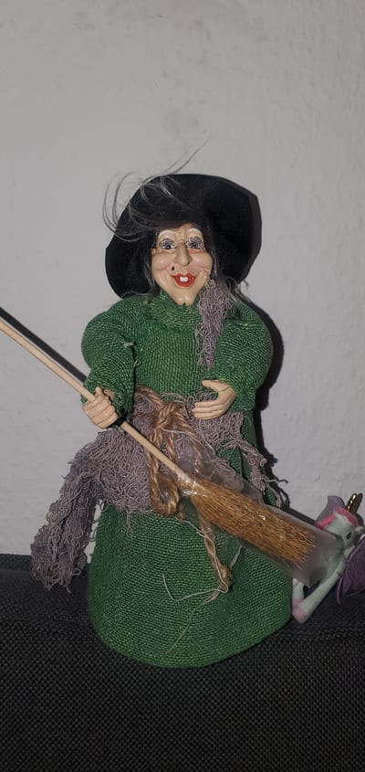 A doll resembling a witch stands in front of a plain white wall, its body draped in a green fabric garment. The witch wears a black hat and a purple scarf. A small broom is held in its hands, attached to a larger object that appears to be a unicorn figure. The background is a white wall, and the foreground is a dark, textured surface. There is no indication of the doll's location in Sasbach, Germany.