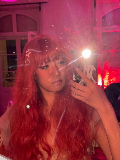 A person with long, wavy, red hair poses, reflected in a mirror. The person wears star-shaped hair accessories near the temple. One hand holds a phone with a black and white patterned case, capturing the image. The reflection reveals a window and pink lighting in the background, with a red light source in the upper portion of the frame. The reflective surface creates a soft, diffused effect, with visible imperfections and light flares.