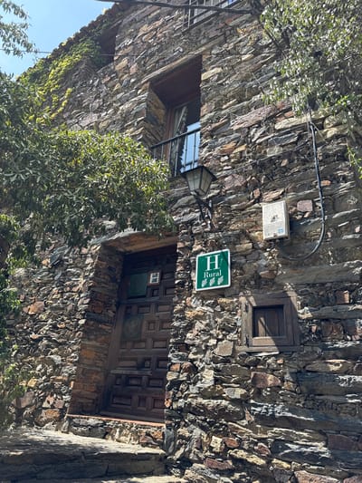 The image shows the exterior of a rustic stone building in Patones De Arriba, Spain. The building features a traditional wooden door and a small window. A green sign with a white 'H' and the word 'Rural' indicates it is a rural accommodation. There are also an outdoor lamp and an electrical box mounted on the stone wall. Lush green foliage climbs the wall and hangs from the roofline, suggesting a natural and somewhat wild environment. The building materials and architectural style are consistent with the characteristic appearance of Patones De Arriba, a village known for its well-preserved slate architecture.